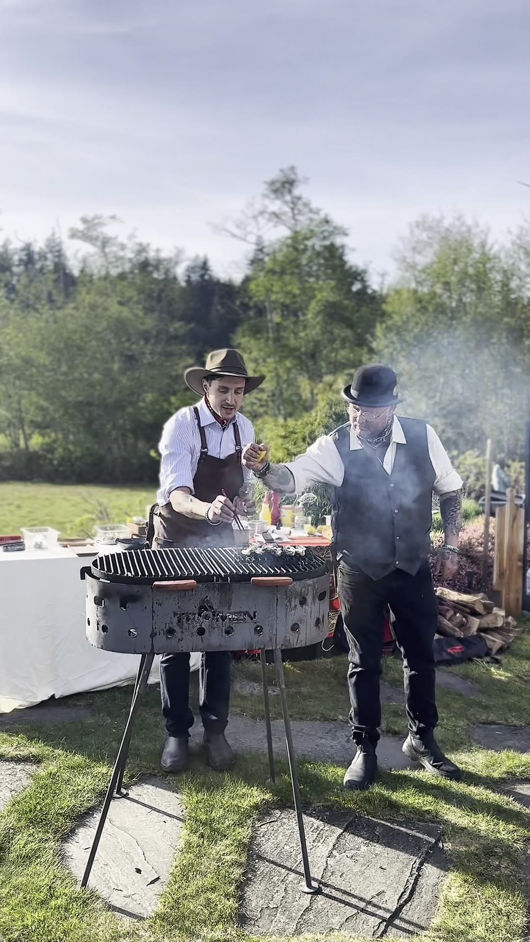 Chef in a hat cooking at an outdoor fire while guests look on